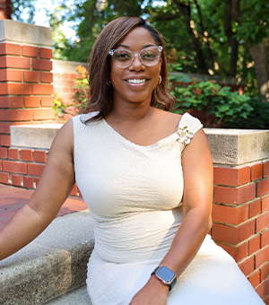 a person sitting on a stone bench in a garden area wearing glasses and a light-colored, one-shoulder dress.